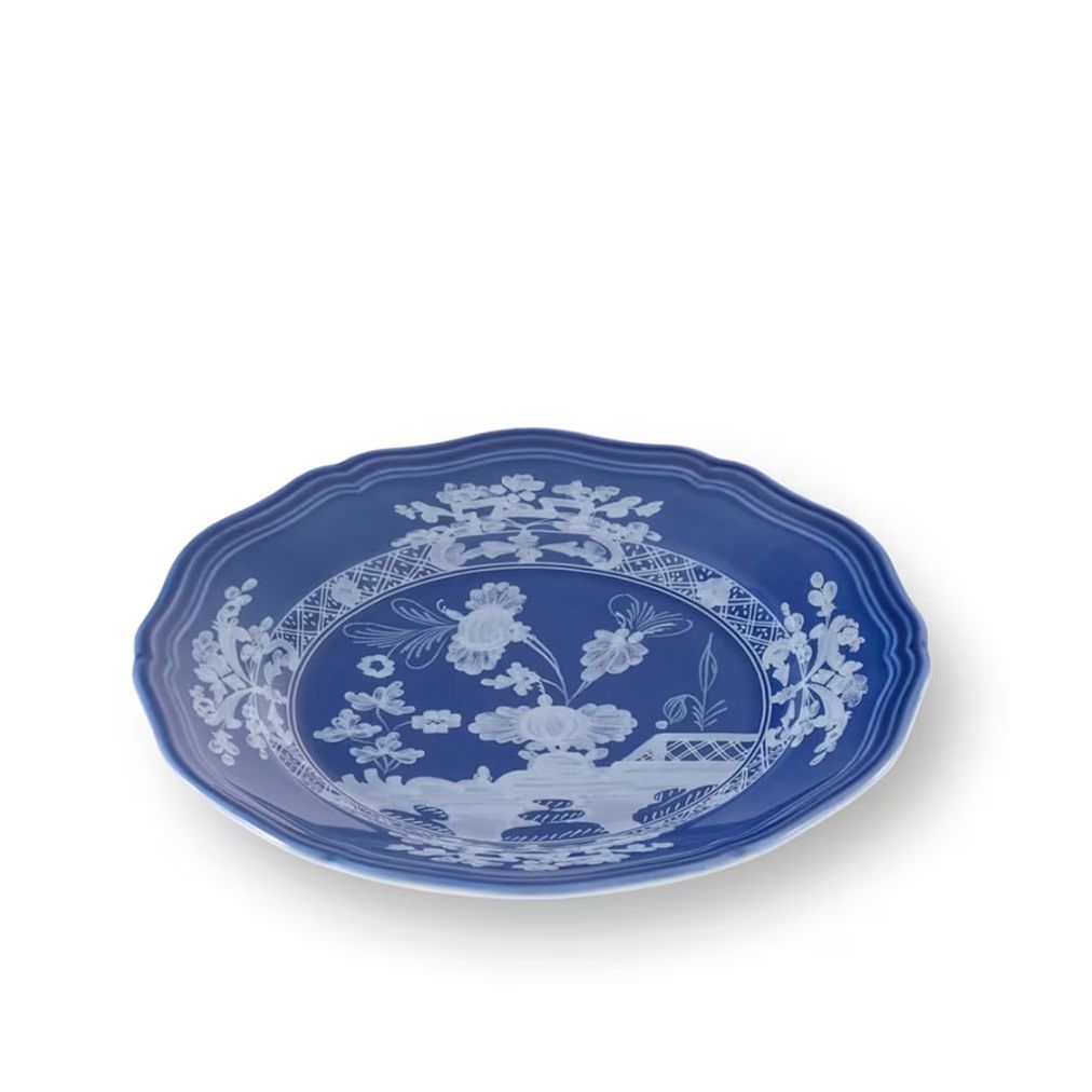 A blue and white ceramic plate with a scalloped rim, featuring a detailed white landscape and floral pattern on a solid blue background, set against a plain white studio backdrop.