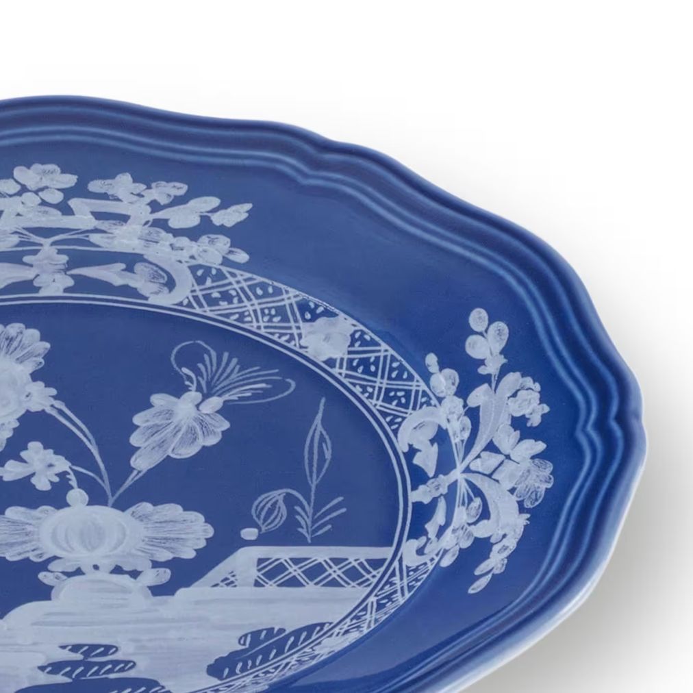 A close-up of a blue ceramic plate with a scalloped rim, featuring a white floral and decorative border pattern on a white background.