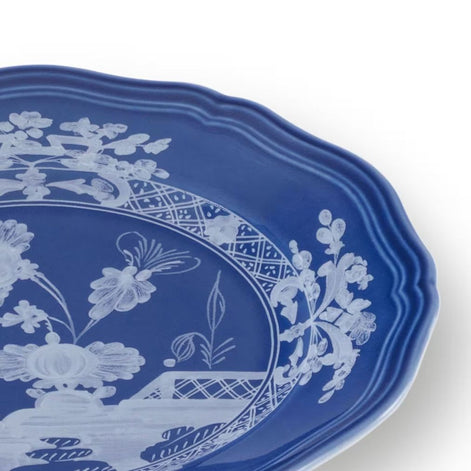 A close-up of a blue ceramic plate with a scalloped rim, featuring a white floral and decorative border pattern on a white background.