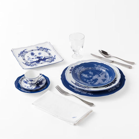 A blue and white porcelain tableware set arranged on a white background, including plates, a cup and saucer, a square plate, a folded napkin, and silver cutlery with a clear drinking glass.