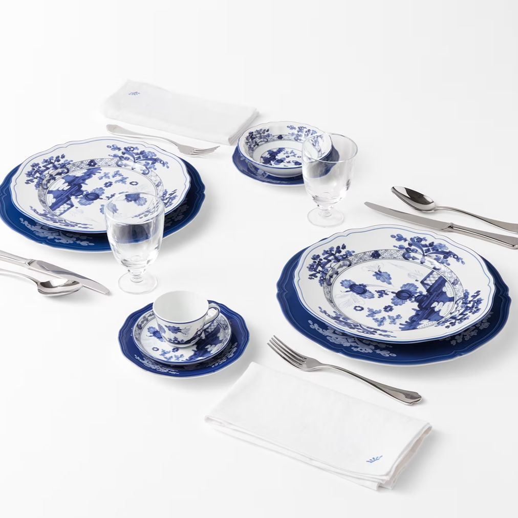 A place setting featuring blue and white porcelain dinnerware, including plates, a teacup and saucer, folded white napkins, and silver flatware, arranged on a clean white background.