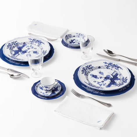A place setting featuring blue and white porcelain dinnerware, including plates, a teacup and saucer, folded white napkins, and silver flatware, arranged on a clean white background.