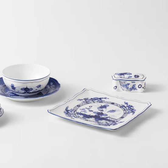 Blue and white porcelain dinnerware set on a light gray background, including a bowl on a saucer, a square plate, and a small lidded container.