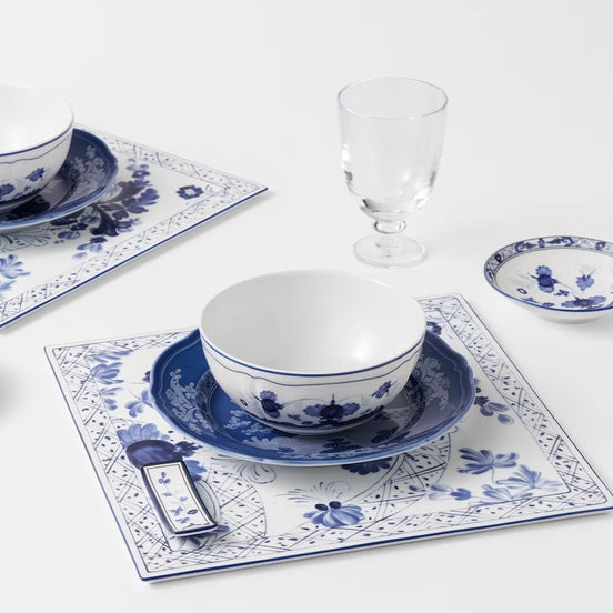 Blue and white porcelain dinnerware set arranged on a white surface, including a bowl, matching saucer, square placemat, small plate, and a clear stemmed glass.