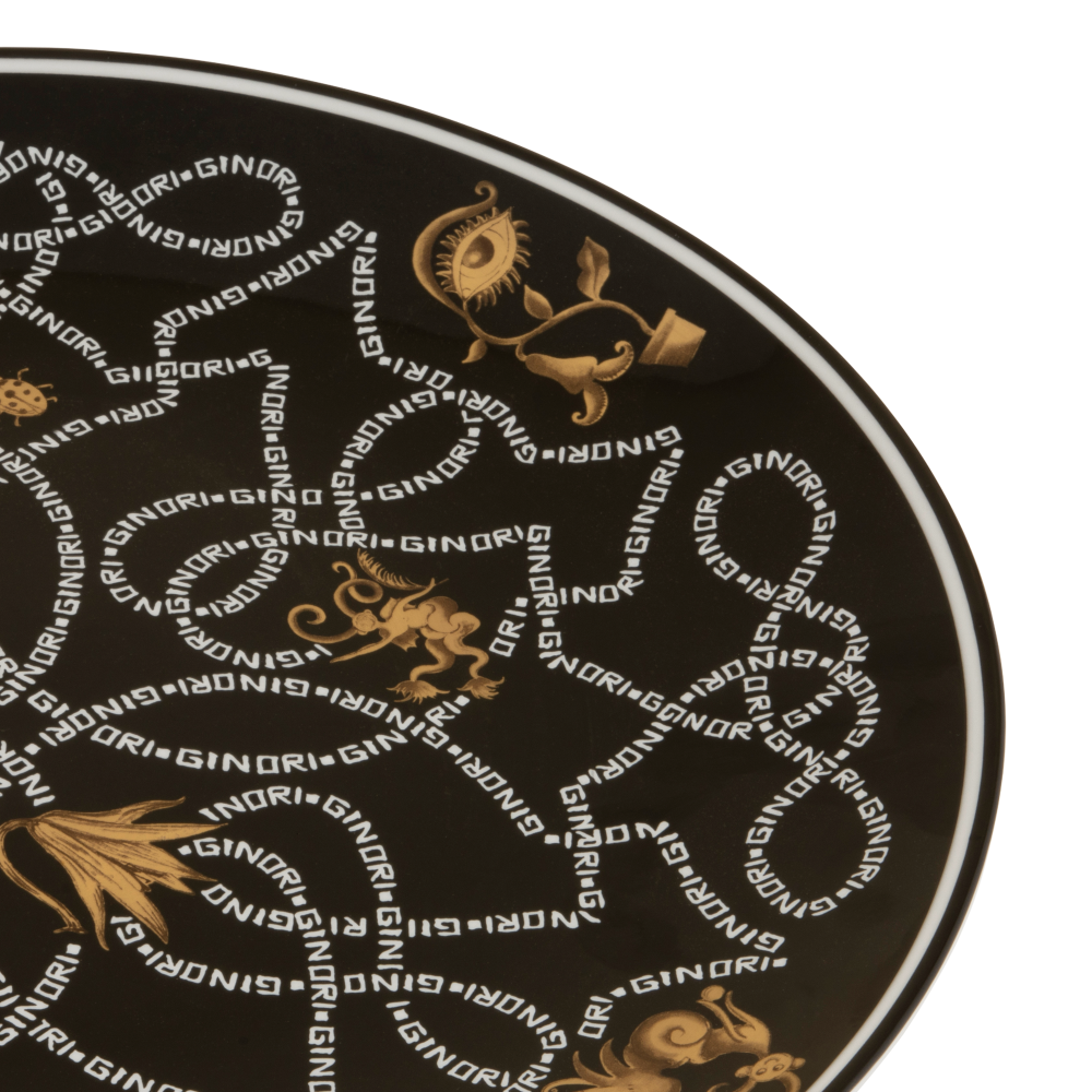 A close-up of a round black plate or platter featuring a repeating gold and white 'SINDRI' monogram pattern with ornate classical motifs.
