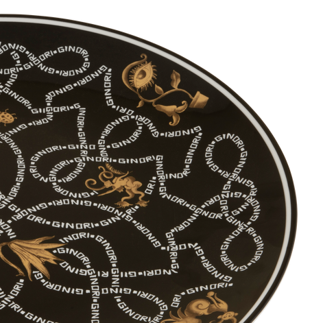 A close-up of a round black plate or platter featuring a repeating gold and white 'SINDRI' monogram pattern with ornate classical motifs.