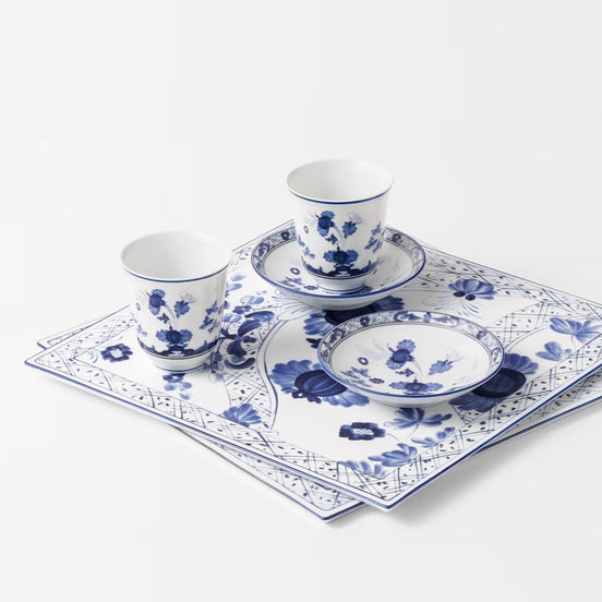 Blue and white porcelain dinnerware set including two cups on saucers, one plate, and matching square placemats with a floral and geometric pattern, arranged on a white background.