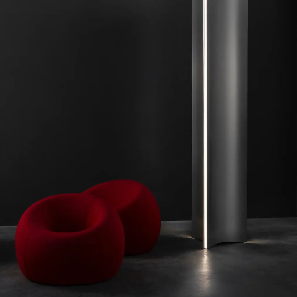 Terzani Stanley floor lamp beside two red sculptural chairs, showing its brushed stainless steel body and illuminated vertical diffuser against a dark wall.