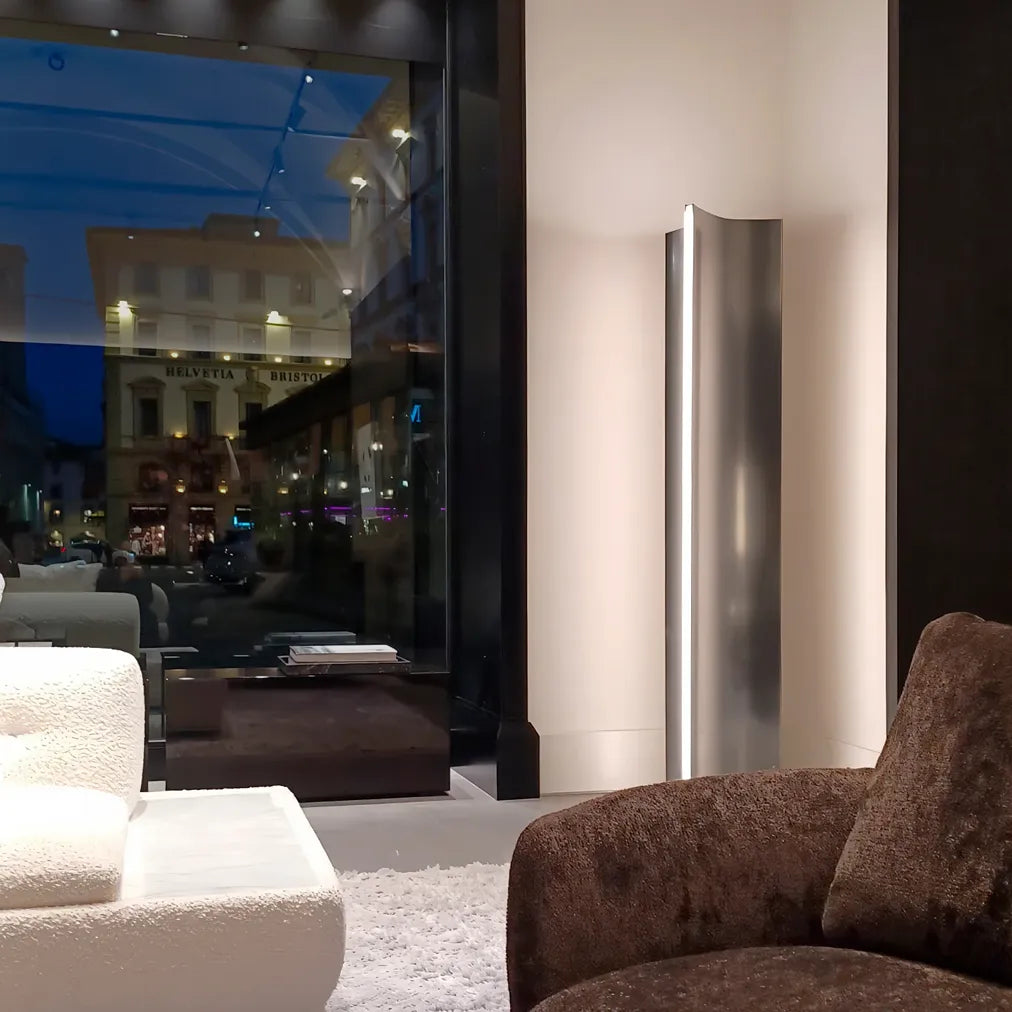 Illuminated Terzani Stanley floor lamp in a contemporary seating area beside a large window, with a curved stainless steel body and vertical blade of light.