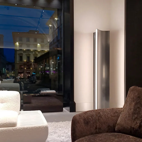 Illuminated Terzani Stanley floor lamp in a contemporary seating area beside a large window, with a curved stainless steel body and vertical blade of light.