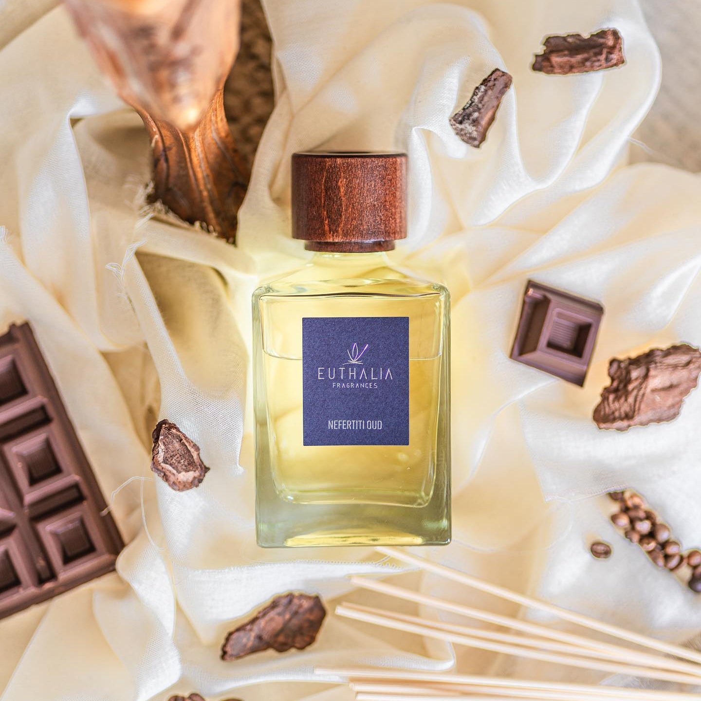 A bottle of Eutelia Fragrances 'Nefertiti Oud' perfume with a dark wood cap and blue label, styled on draped cream fabric with chocolate pieces and coffee beans. At Archideco Interior Gallery.