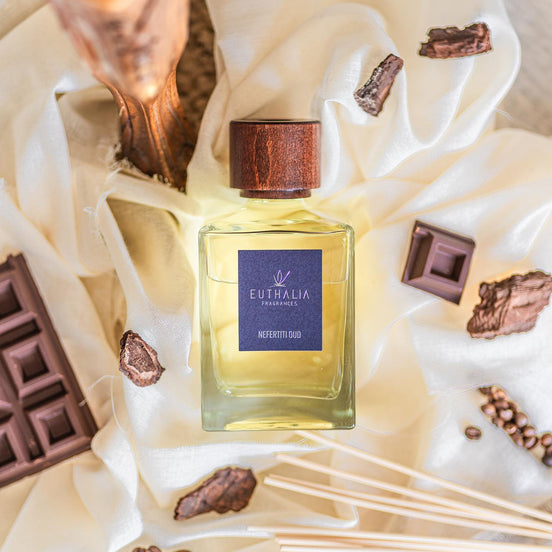 A bottle of Eutelia Fragrances 'Nefertiti Oud' perfume with a dark wood cap and blue label, styled on draped cream fabric with chocolate pieces and coffee beans. At Archideco Interior Gallery.