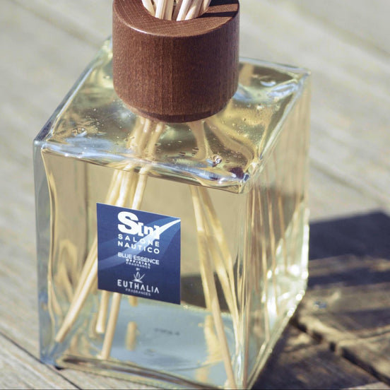 A reed diffuser bottle with a square clear glass container, pale liquid, visible reeds, a dark brown wooden stopper, and a blue label reading 'Salone Nautico Blue Essence' by Euthalia Fragrances.