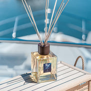 A square glass reed diffuser bottle filled with pale yellow liquid, topped with a dark brown cap and natural wood reeds, placed on a light wooden surface with a blurred blue water background. Euthalia Fragrances at Archideco Interior Gallery