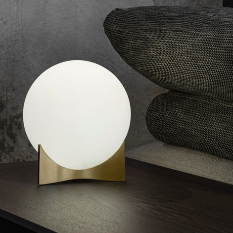 Terzani Oscar table lamp with a glowing opal glass globe and gold-tone base on a dark console beside textured cushions.