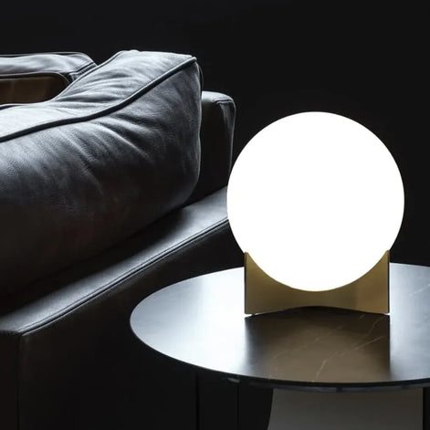 Illuminated Terzani Oscar table lamp with a gold-tone base on a round dark side table beside a black leather sofa.