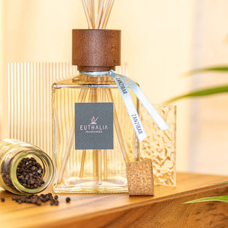 A clear glass reed diffuser bottle with a brown wooden top and reeds, tied with a white ribbon labeled EUTHALIA AROMAS, displayed on a wooden surface with a small jar of peppercorns and a textured glass in a warm indoor setting.