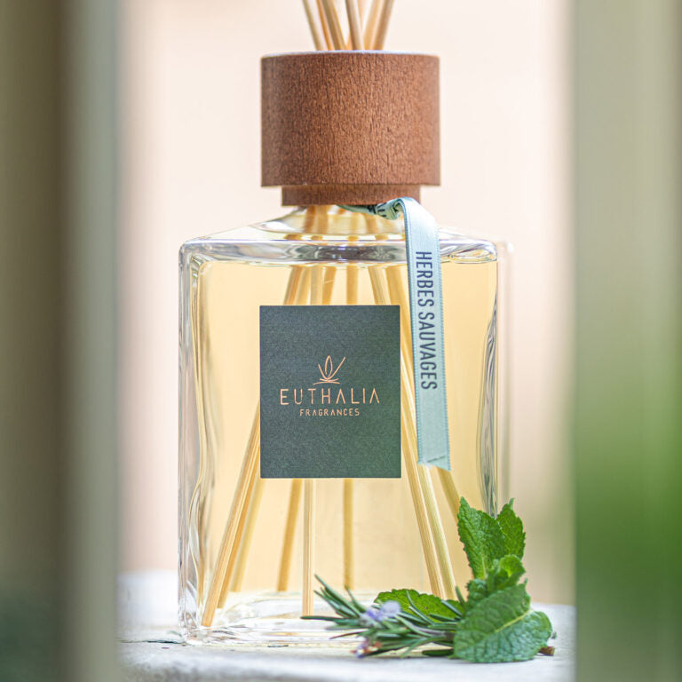 A clear glass reed diffuser bottle filled with pale golden liquid, topped with a brown cork-like reed cap and natural reeds. A dark green square label reads EUTHALIA FRAGRANCES, with a light blue ribbon tag that says HERBES SAUVAGES. At Archideco Interior Gallery.